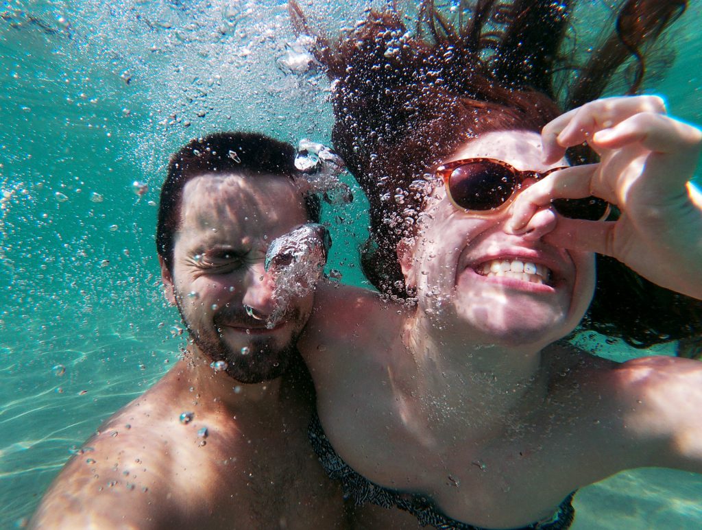 two people underwater with bubbles all around them. One is pinching her nose with her fingers and wearing sunglasses as her hair floats in the clear, turquoise water