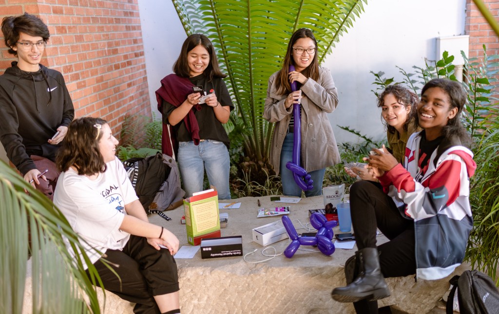 a group of 6 students sit on a large stone in the School of Art, Art Gallery Courtyard at Long Beach State University and play games and eat snacks.