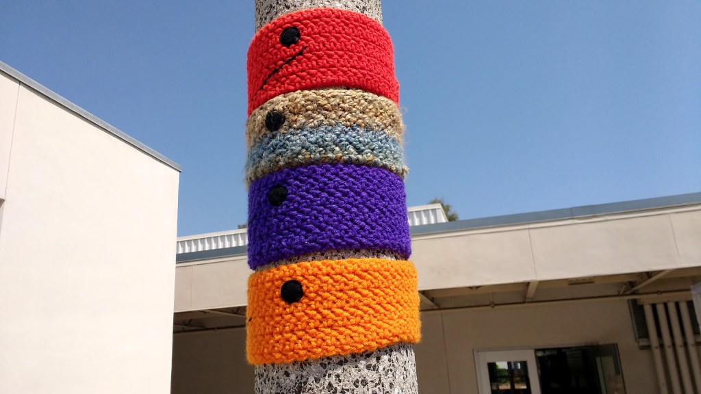 4 "collars" in red, brown, purple, and orange yarn are woven around a light pole at the CSULB, College of the Arts, School of Art, near the Art Gallery Complex
